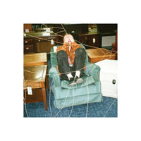 A person reclining in a dusty green armchair inside a furniture store, surrounded by other chairs, wooden tables, and a white storage cabinet. The photo has a web-like overlay pattern across the frame.