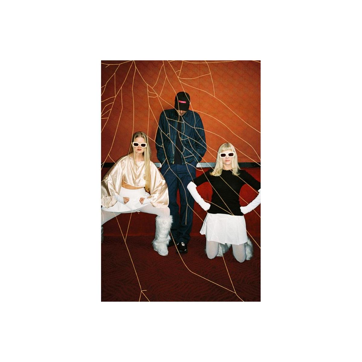 Three models pose in a stylized studio setup with a textured orange-brown wall, gold web-like lines, and a dark patterned floor. One model wears a shiny cream satin top and white pants, seated on a bench.