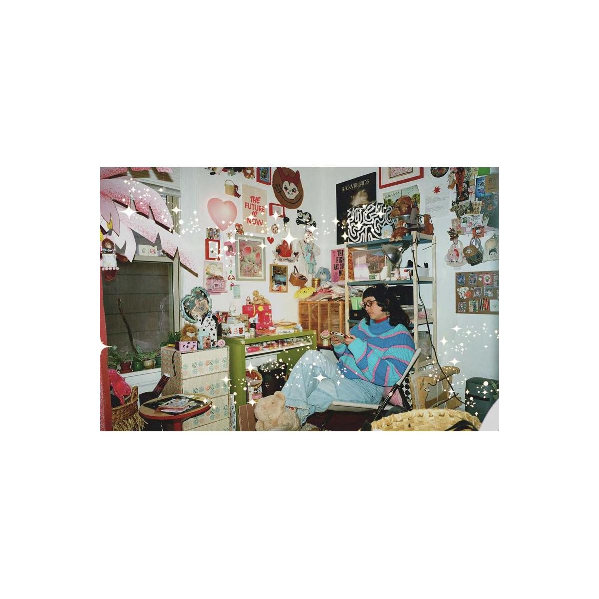 A cozy, cluttered room decorated with pink and red heart motifs, posters, and shelves, featuring a person sitting in a chair wearing a colorful striped sweater and light blue jeans.