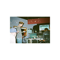 A candid photo inside a recording studio with audio equipment, speakers, and computer monitors. A man in a gray t-shirt and jeans stands holding a camera, while another man sits at a desk facing a monitor.