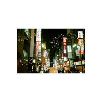 A nighttime street scene in a Japanese city with glowing multicolored neon signs, storefront signs in Japanese, and many blurred pedestrians walking down the street. The image has a warm vintage filter with soft focus and decorative starburst overlays.