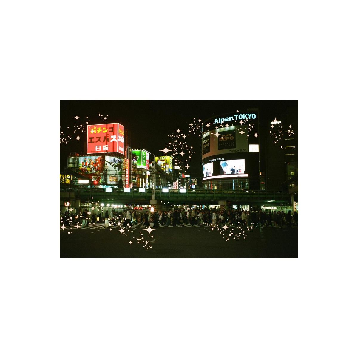 A nighttime city scene with brightly lit billboards and signs, including visible text such as AlpenTokyo and Japanese signage, with a crowd of people crossing a street in the foreground. The image has decorative sparkle/star overlays.