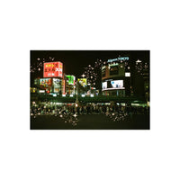 A nighttime city scene with brightly lit billboards and signs, including visible text such as AlpenTokyo and Japanese signage, with a crowd of people crossing a street in the foreground. The image has decorative sparkle/star overlays.