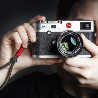 A person is holding a camera up to their face, with the camera's wrist strap attached to their wrist. The camera has a black color and a classic design.