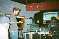 A candid photo inside a recording studio with audio equipment, speakers, and computer monitors. A man in a gray t-shirt and jeans stands holding a camera, while another man sits at a desk facing a monitor.