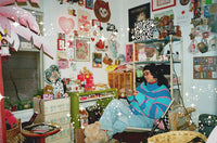 A cozy, cluttered room decorated with pink and red heart motifs, posters, and shelves, featuring a person sitting in a chair wearing a colorful striped sweater and light blue jeans.