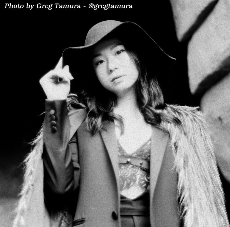 Black and white portrait of a woman wearing a wide-brimmed hat, a patterned top, and a textured jacket with a shawl collar, posed against a dark background. Text at the top reads 'Photo by Greg Tamura - @gregtamura'.