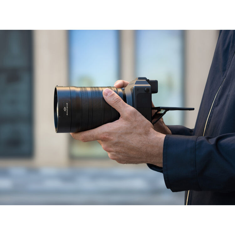 A close-up of a person holding a black camera with a large telephoto lens, shown from the side against a blurred outdoor background.