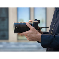 A close-up of a person holding a black camera with a large telephoto lens, shown from the side against a blurred outdoor background.