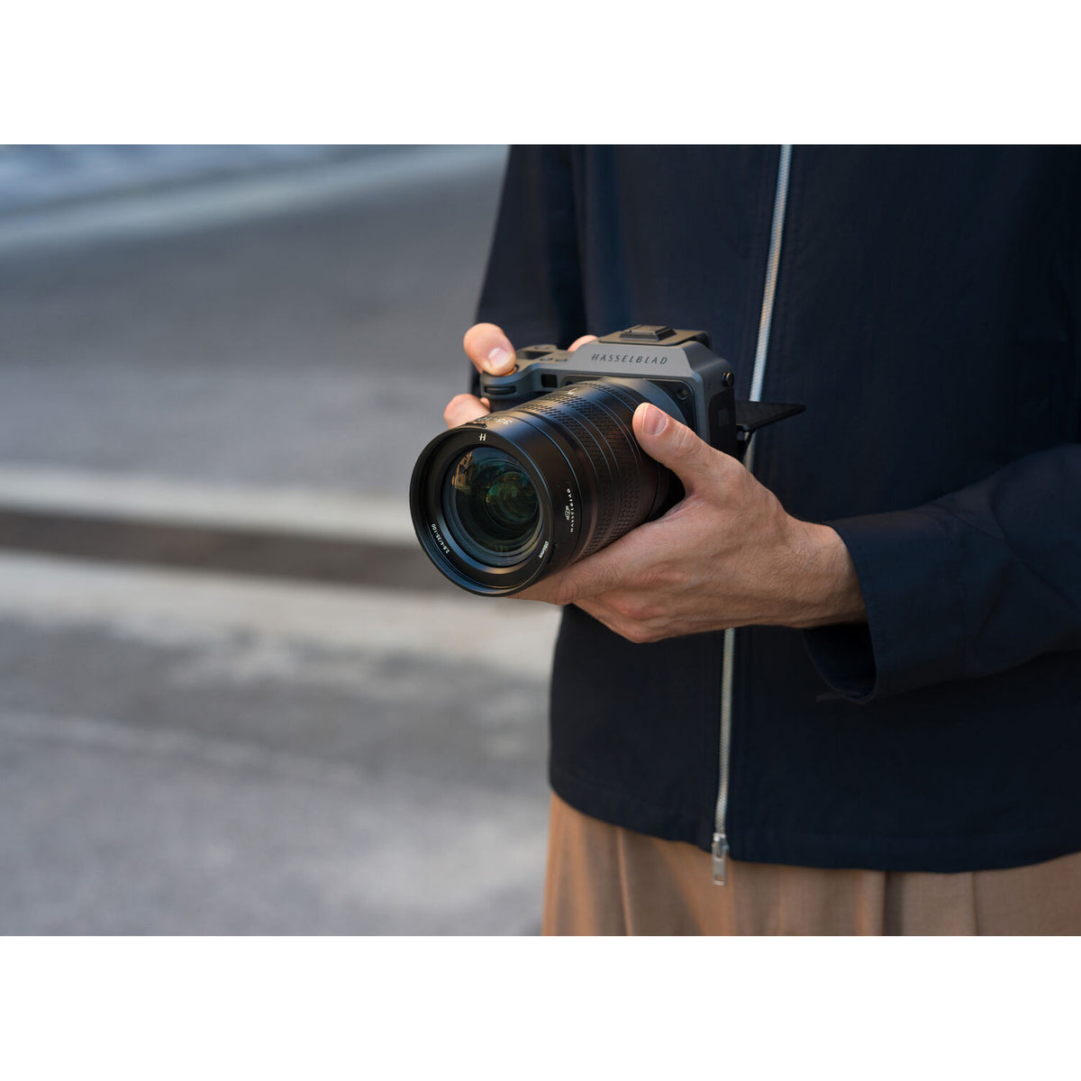 A close-up of a person holding a Sony Alpha SLR camera with a large lens, wearing a dark navy jacket and beige pants against an outdoor blurred background.