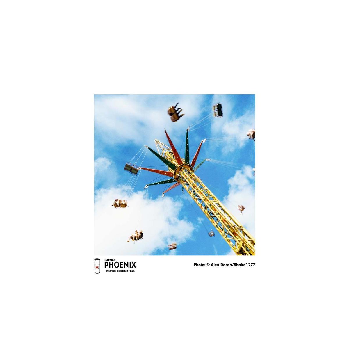 A square photographic print of an amusement park swing ride against a blue sky with clouds, shown centered on a white background with a small PHOENIX logo and photo credit at the bottom.