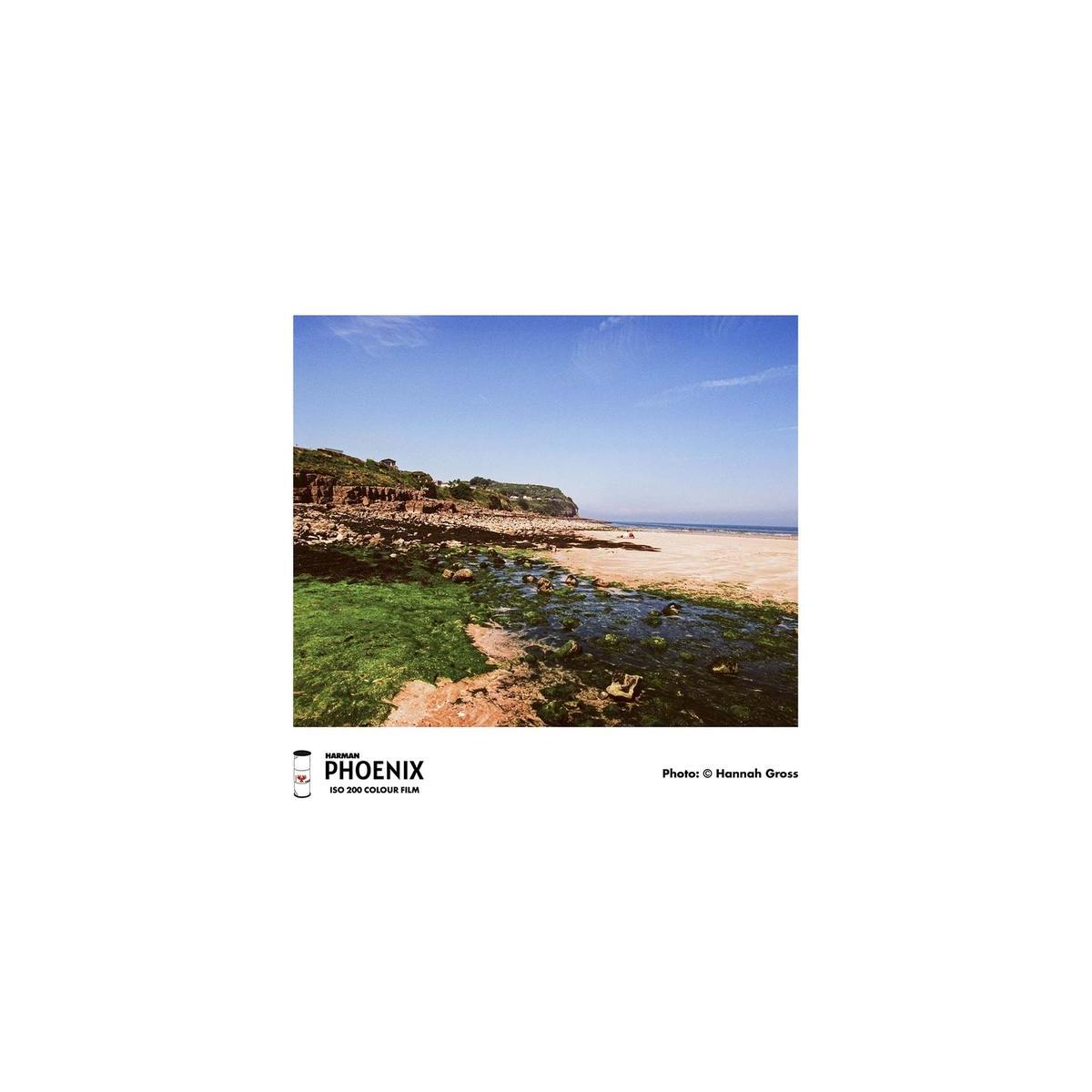 A square photographic print showing a coastal shoreline with rocky tide pools, green seaweed-covered rocks, a pale sandy beach, and blue sky, presented on a white page with the text 'PHOENIX ISO 200 COLOUR FILM' and 'Photo: Hannah Gross'.