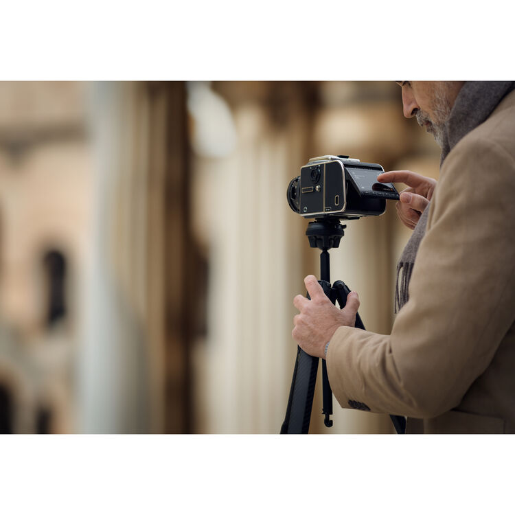 A man in a beige coat adjusts a black camera mounted on a black tripod, photographed in a softly blurred outdoor setting.