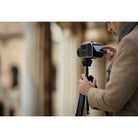 A man in a beige coat adjusts a black camera mounted on a black tripod, photographed in a softly blurred outdoor setting.