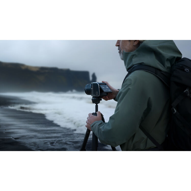 A man in a green hooded jacket and black backpack stands on a dark beach holding a camera on a tripod, with ocean waves and cliffs in the background.