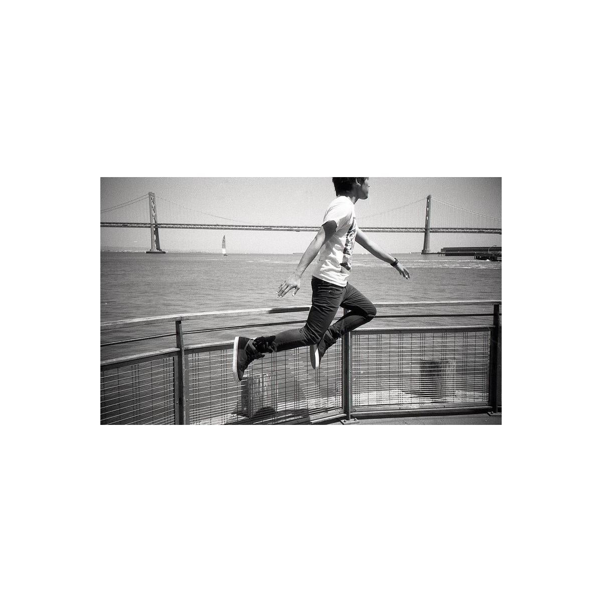 A black and white lifestyle photograph of a person mid-jump over a metal railing, with a large suspension bridge and water in the background.