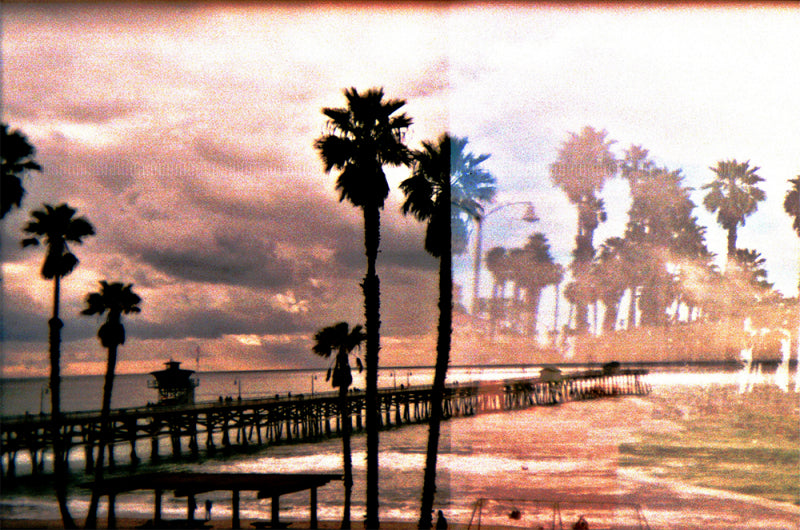 Double exposure film photo of a beach pier and palm trees with dreamy, lo-fi colors and nostalgic vignetting.