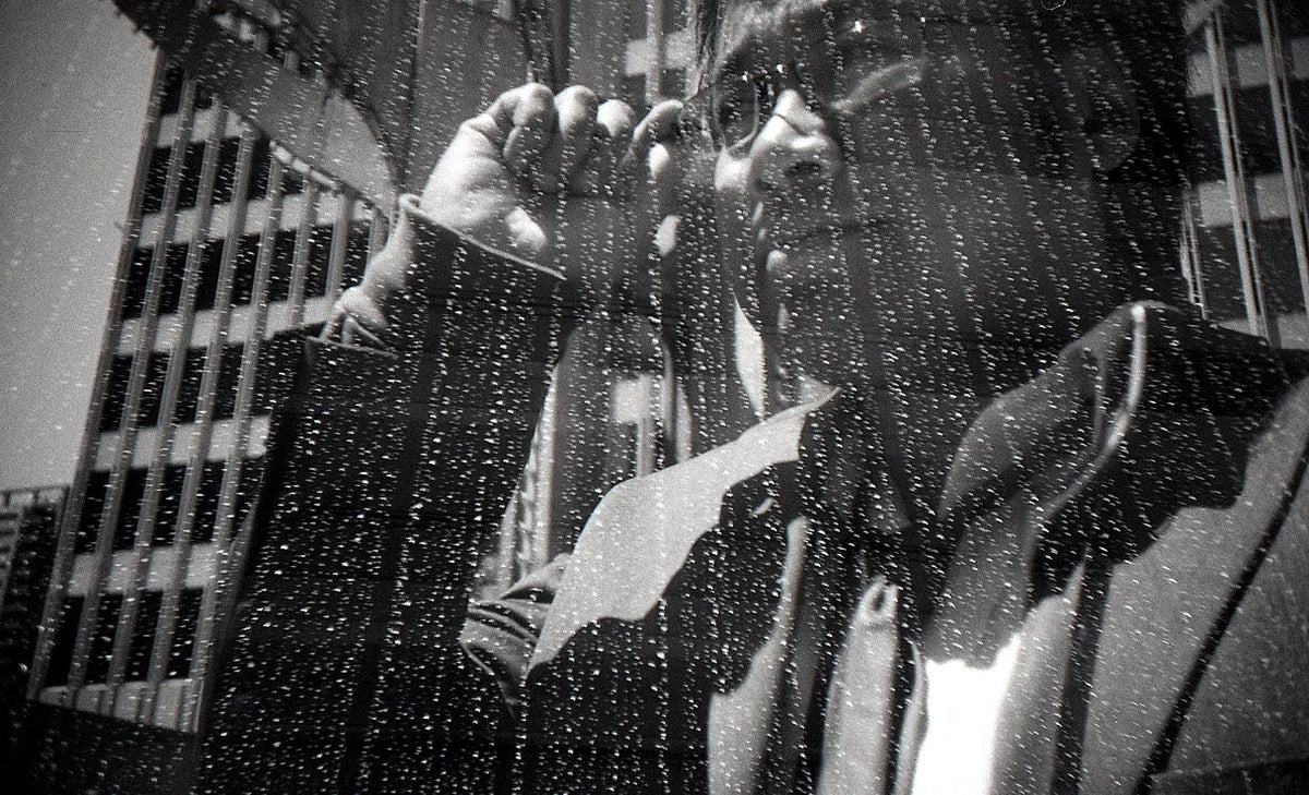 Black and white photograph of a person in a raincoat and glasses looking through binoculars, with raindrops on a window and a city building in the background.
