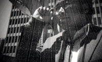 Black and white photograph of a person in a raincoat and glasses looking through binoculars, with raindrops on a window and a city building in the background.