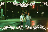 Three people standing at night outside a convenience store under green and red store lights, with a white tribal graphic border overlaying the top and bottom of the photo.