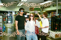 Three people standing in a convenience store aisle, with a decorative translucent tribal pattern overlaid across the entire image. The person on the left wears a black t-shirt and jeans.