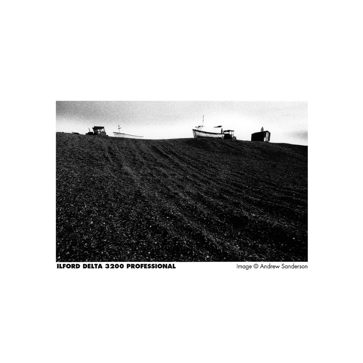 A black and white fine art photograph print of a landscape, featuring boats on a dark, textured hillside under a pale sky. The print is presented with a white border containing the text 'ILFORD DELTA 3200 PROFESSIONAL' and 'Image © Andrew Sanderson'.
