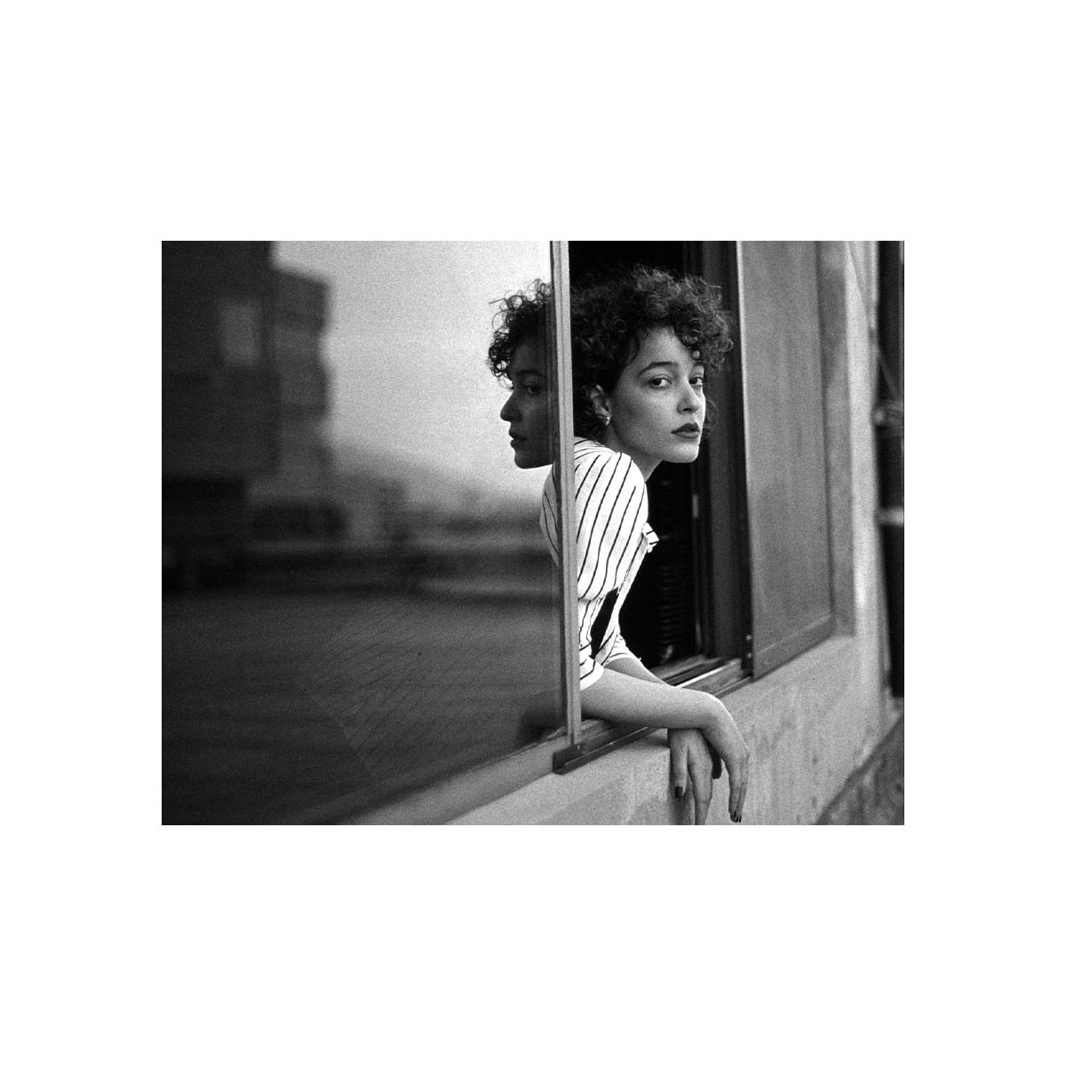 A black and white photograph print featuring a woman with curly hair leaning against a window frame, looking off-camera. The print is centered on a solid white background.