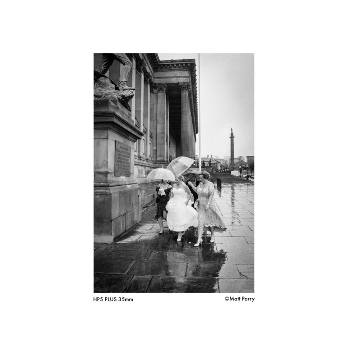 A black and white photographic art print titled 'HPS PLUS 35mm' by Math Parry, depicting a rainy street scene with women holding umbrellas next to a classical stone building.