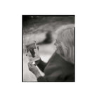 A black and white photographic print featuring an elderly woman holding a small photograph in her hands, shown against a white border.