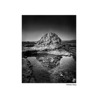 A black and white fine art photograph print of a rugged rock formation reflected in a shallow pool of water, presented with a white border and the photographer's name, Natalie Oberg, printed in the bottom right corner.