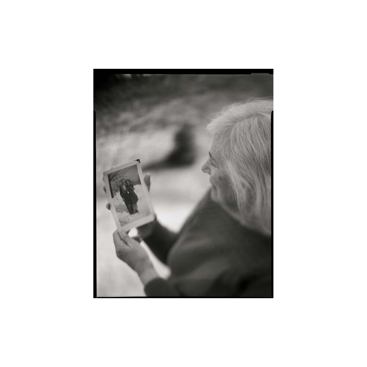 A black and white square photograph print featuring an elderly woman in profile holding up a small photograph of two people standing together, set against a plain white background.