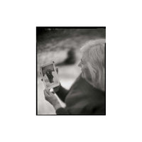 A black and white square photograph print featuring an elderly woman in profile holding up a small photograph of two people standing together, set against a plain white background.