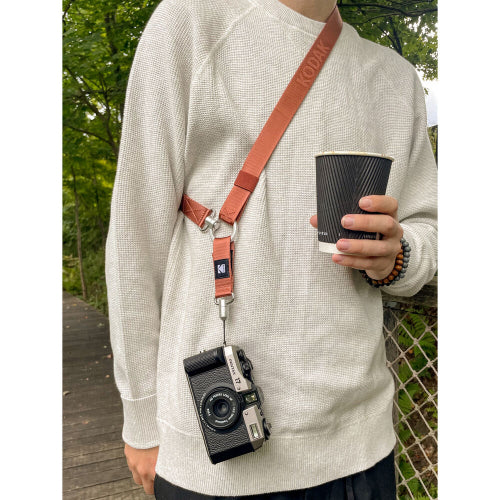 A person wearing a light beige knit sweater carries a brown leather camera strap across their chest, holding a black coffee cup. The strap is attached to a vintage black and silver film camera hanging from a silver metal clip.