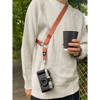 A person wearing a light beige knit sweater carries a brown leather camera strap across their chest, holding a black coffee cup. The strap is attached to a vintage black and silver film camera hanging from a silver metal clip.