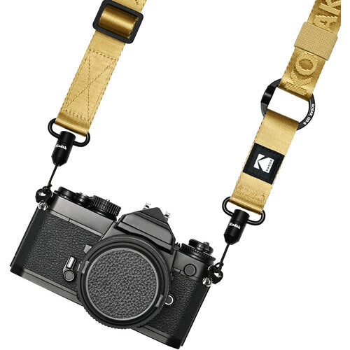 A black film camera body with a tan webbed camera strap attached. The strap features a black logo patch with the Kodak 'K' logo and the word 'KODAK' printed on it, along with black metal quick-release clips.