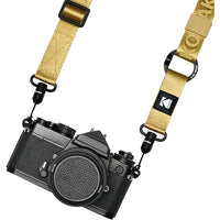 A black film camera body with a tan webbed camera strap attached. The strap features a black logo patch with the Kodak 'K' logo and the word 'KODAK' printed on it, along with black metal quick-release clips.