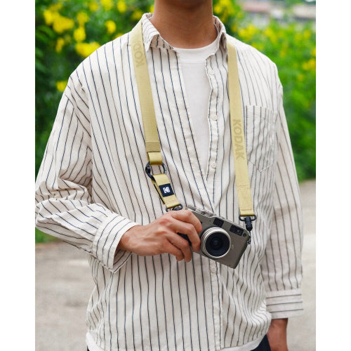 A beige Kodak camera strap worn cross-body over a cream and navy striped button-up shirt, with a person holding a silver film camera in front of a blurred outdoor background of green foliage and yellow flowers.