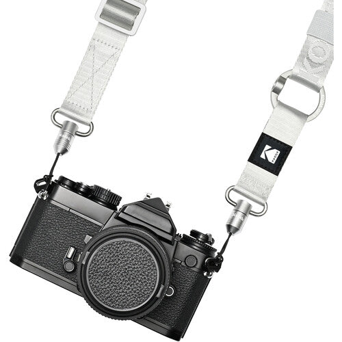 A white woven camera strap with silver metal hardware and a black square patch featuring the Kodak logo, shown attached to a black film camera on a white background.