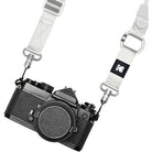A white woven camera strap with silver metal hardware and a black square patch featuring the Kodak logo, shown attached to a black film camera on a white background.