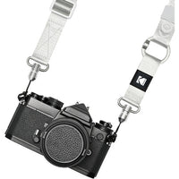 A white woven camera strap with silver metal hardware and a black square patch featuring the Kodak logo, shown attached to a black film camera on a white background.