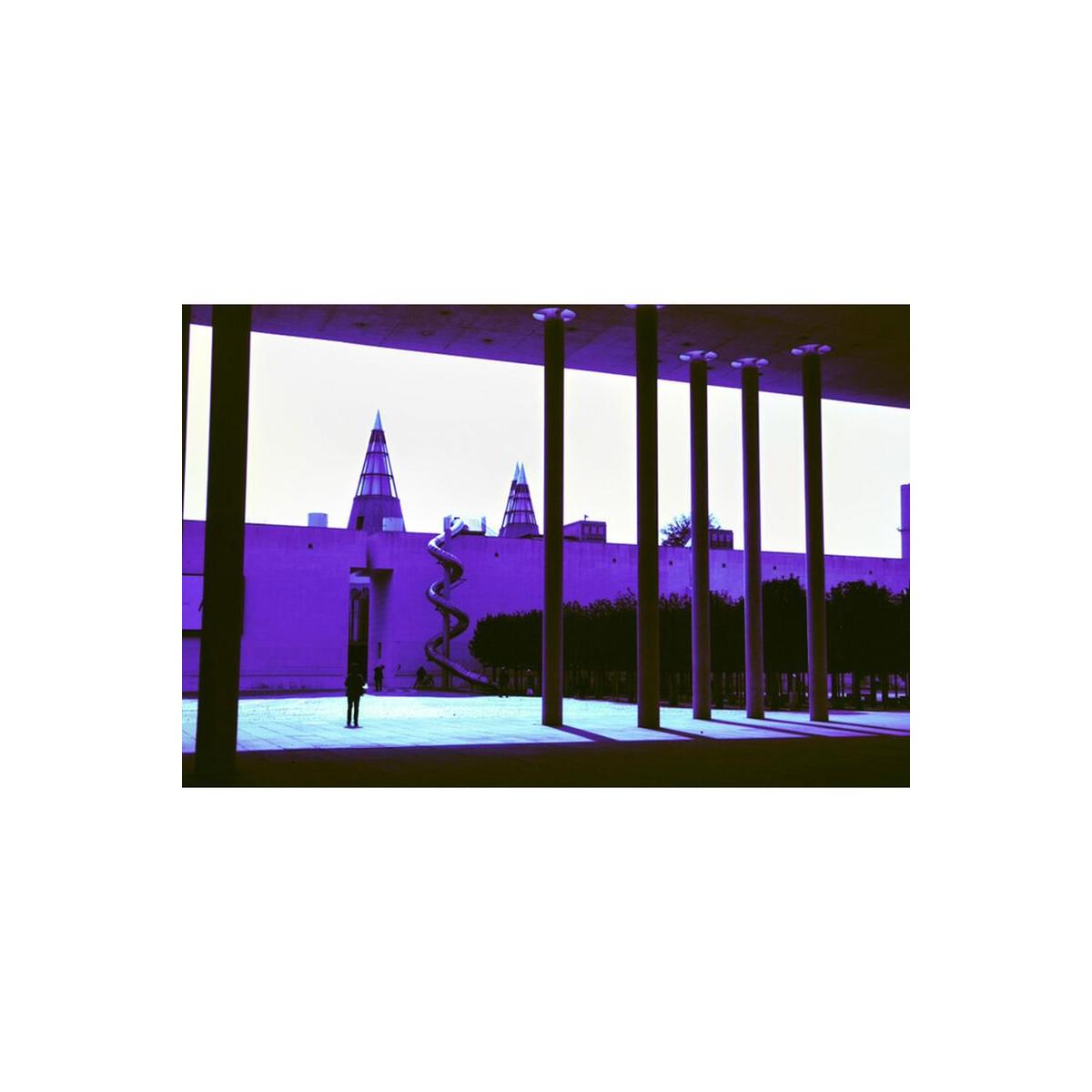 Architectural scene with strong purple and blue color shifts, featuring tall pillars and a spiral slide in bright daylight.