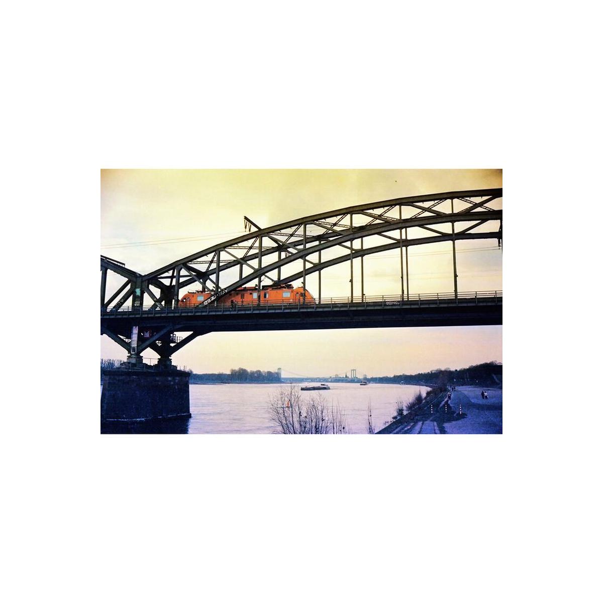 Orange train crossing a steel bridge over a river, captured with KONO! Moonstruck 200 film's muted, experimental color tones.