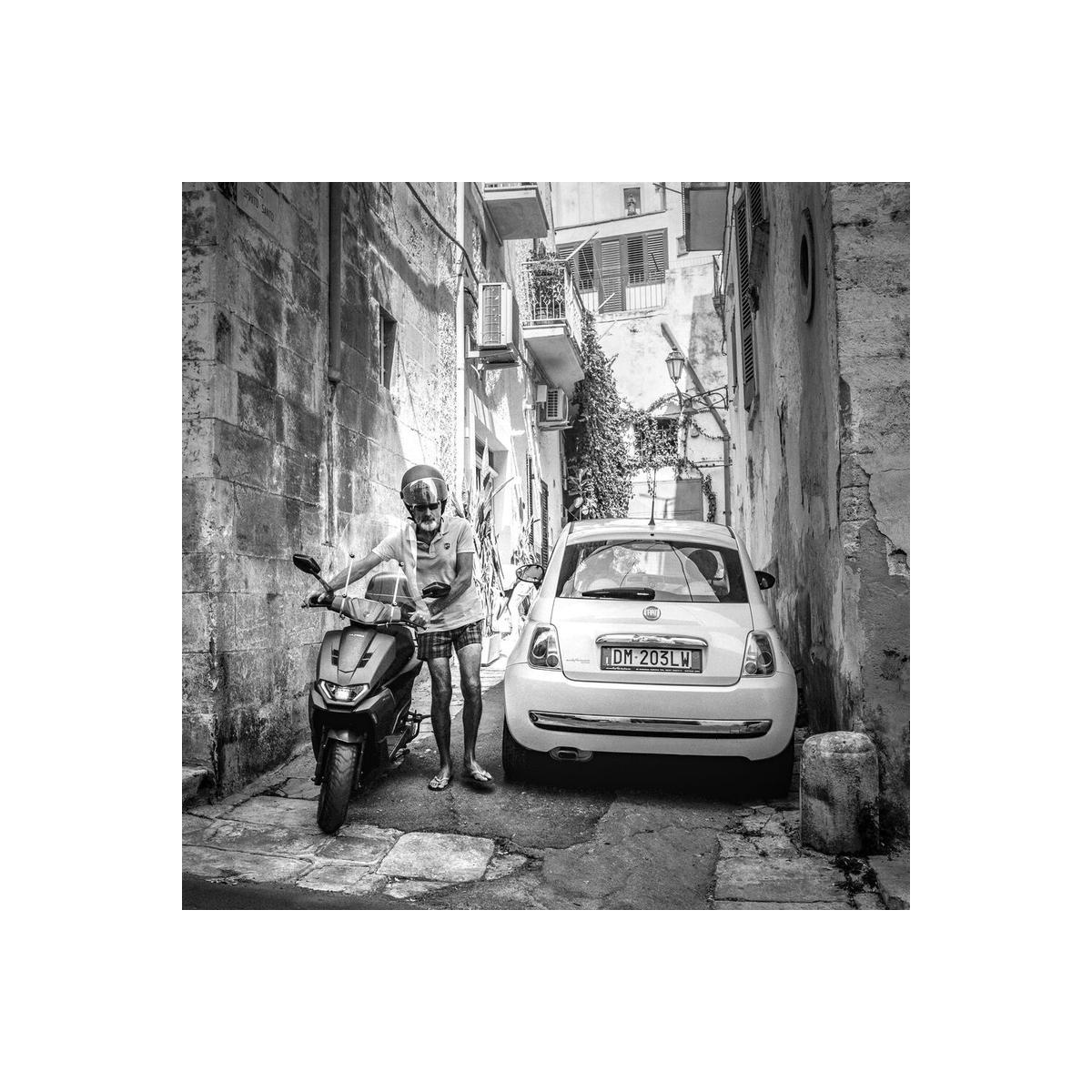 Black and white street scene featuring a man on a scooter next to a parked Fiat in a narrow, sunlit stone alleyway.