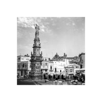 Black and white 35mm film shot of an ornate stone monument in an Italian town square with cafe tables and historic buildings.