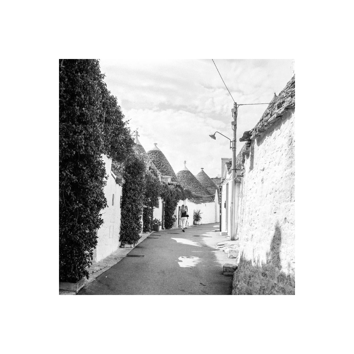 Black and white street scene in Alberobello featuring traditional stone trulli houses and a person walking down the path.