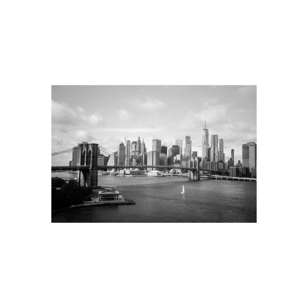 Black and white film photograph of the Brooklyn Bridge and Manhattan skyline with a sailboat on the East River.