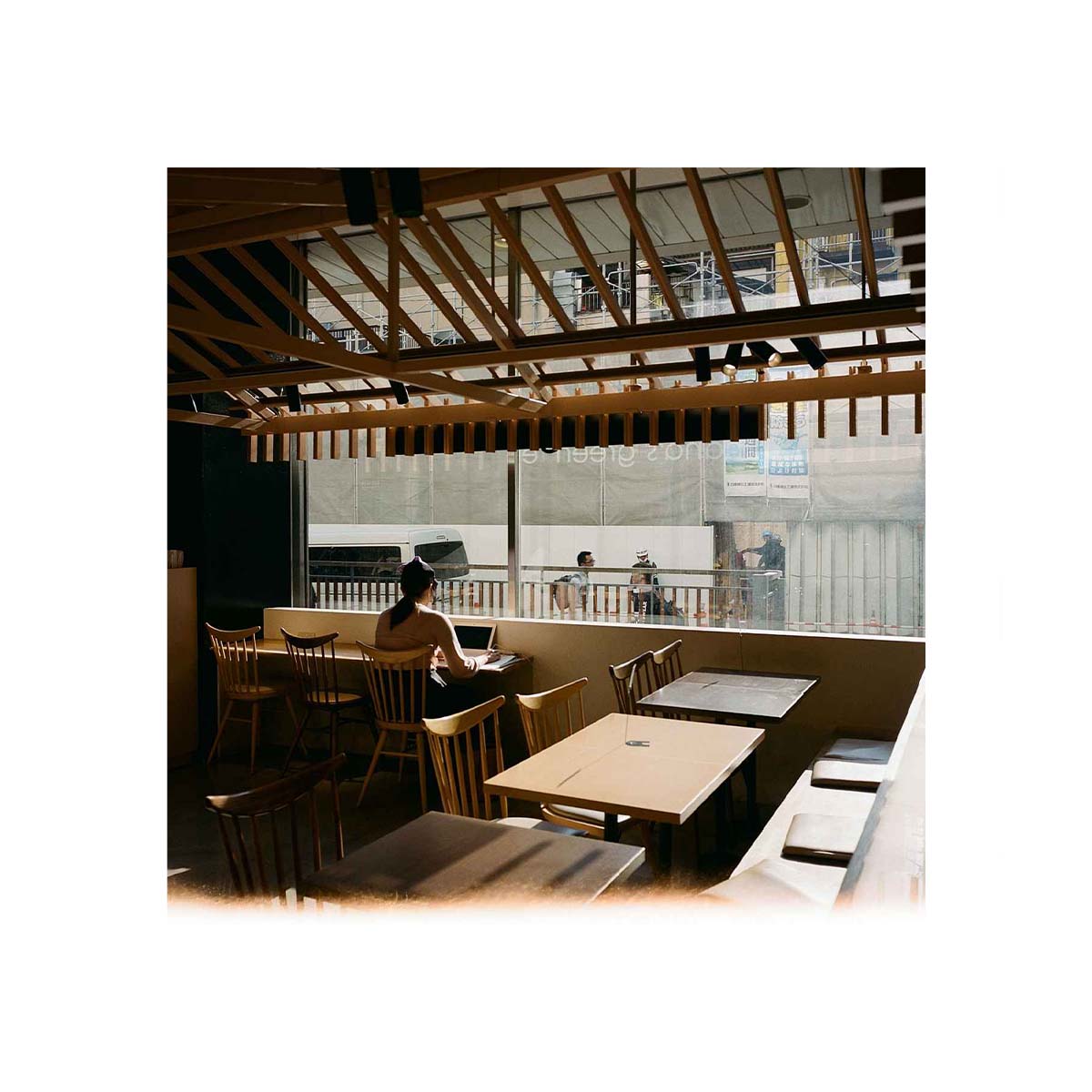 A warm-toned interior scene of a cafe or restaurant with wooden tables and chairs, a person seated at a table, and a large window showing an outdoor construction or balcony area. The photo has a soft, slightly hazy aesthetic.