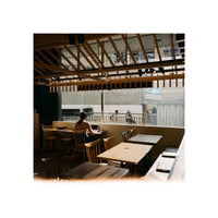 A warm-toned interior scene of a cafe or restaurant with wooden tables and chairs, a person seated at a table, and a large window showing an outdoor construction or balcony area. The photo has a soft, slightly hazy aesthetic.