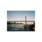 A scenic photograph of a suspension bridge over a river with boats on the water and a city skyline in the background, presented as a print centered on a white background.