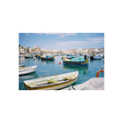 A scenic photograph of a Mediterranean harbor with small boats moored in calm blue water, white buildings along the shoreline, and a bright sky. The image is presented with a wide white border, suggesting it is sold as a photo print or wall art.